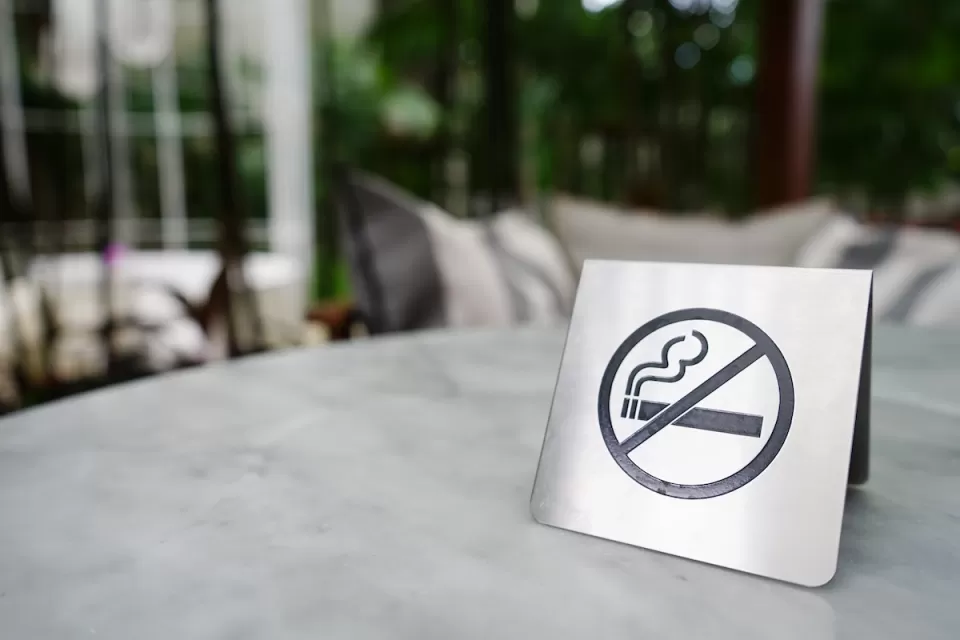 Close-up of silver sign indicating 'no smoking' with cigarette inside circle with line. Sign is sitting on patio table outdoors.